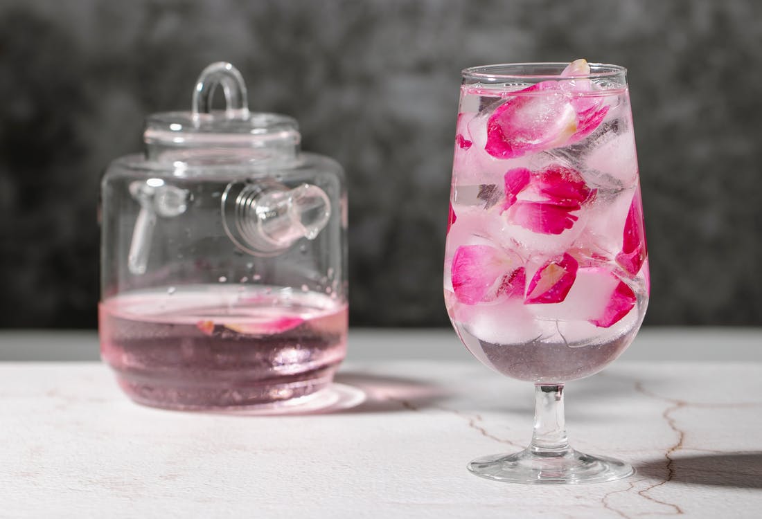Crystal glass full of fresh beverage with pink rose petals and ice cubes place on table near jar with drink